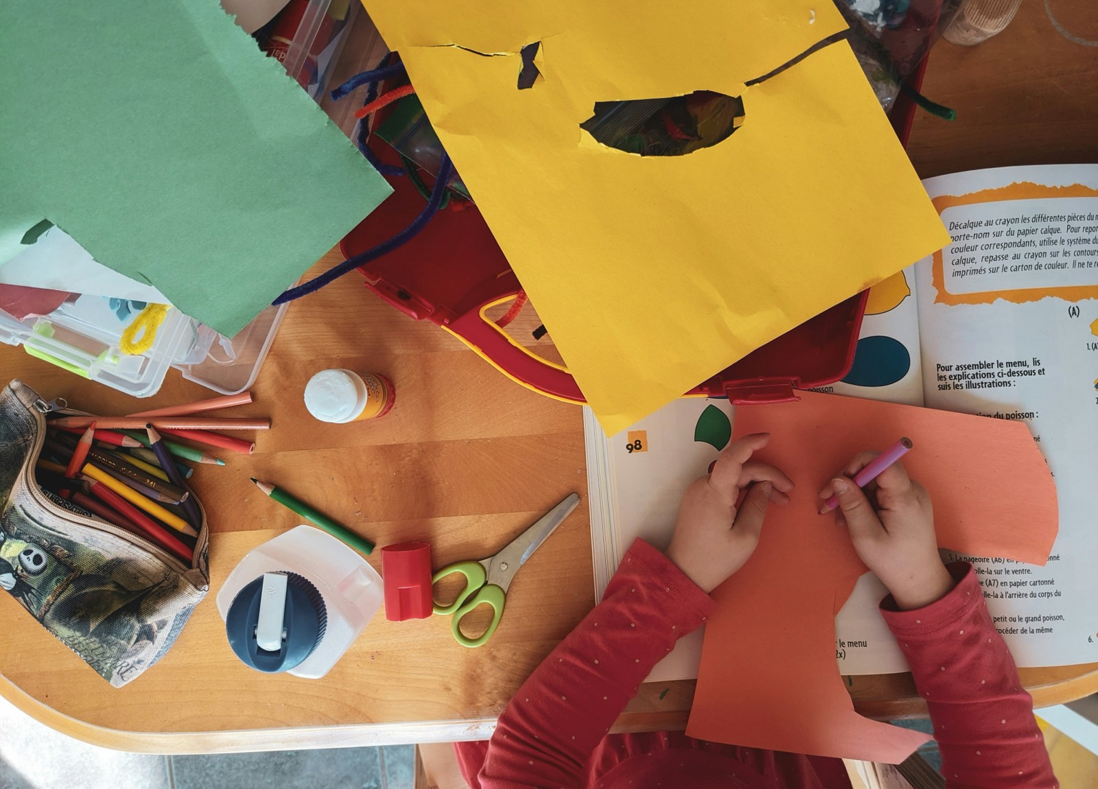 Un enfant dessine au crayon sur une feuille orange, entouré de crayons, de ciseaux et de papiers colorés